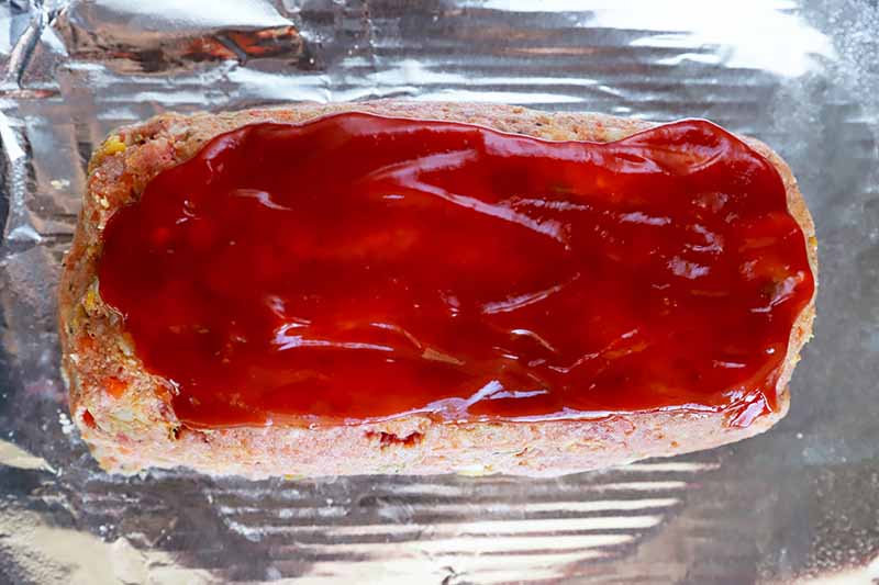 Horizontal image of a glazed unbaked rectangular mound of raw ground beef on a foil lined baking sheet.