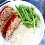 Horizontal top-down image of two slices of glazed meatloaf on a white plate with mashed potatoes and green beans.