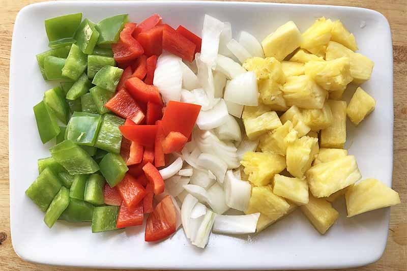 Horizontal image of neat rows of chopped peppers, onions, and pineapple on a white plate.