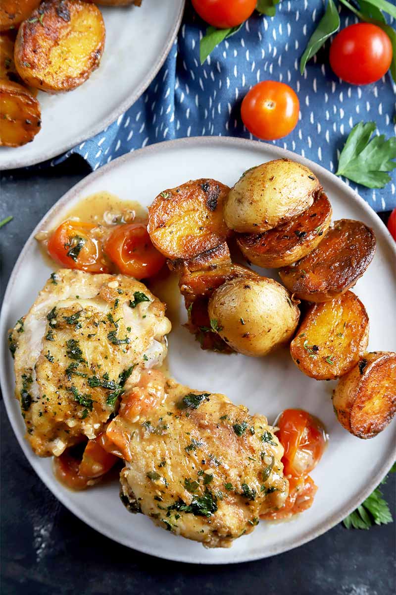Vertical top-down image of a plate with potatoes and a cooked chicken dish seasoned with fresh herbs, next to a a blue towel and fresh tomatoes.