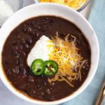 Horizontal image of a white bowl with black bean soup with cheese, sour cream, and jalapeño garnish with a blue napkin and a spoon.