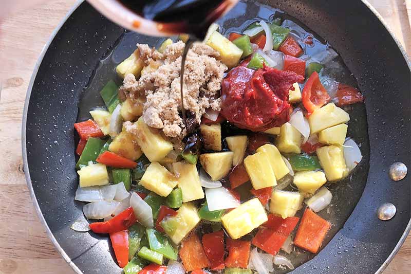Horizontal image of a skillet with chopped onion, pepper, and pineapple with assorted seasonings being poured inside.