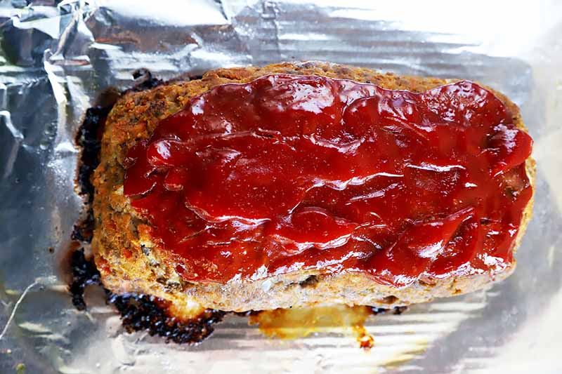 Horizontal image of a baked mound of ground beef with a ketchup glaze on a foil-lined baking sheet.