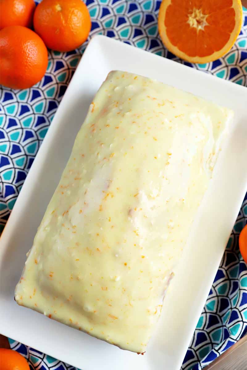 Vertical top-down image of a whole rectangular baked dessert covered in a white glaze on a white plate on top of a pattered blue napkin surrounded by whole citrus fruit.