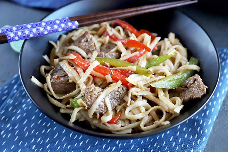 Horizontal image of a black bowl filled with a steak, veggie, and noodle dish with chopsticks and a blue towel.
