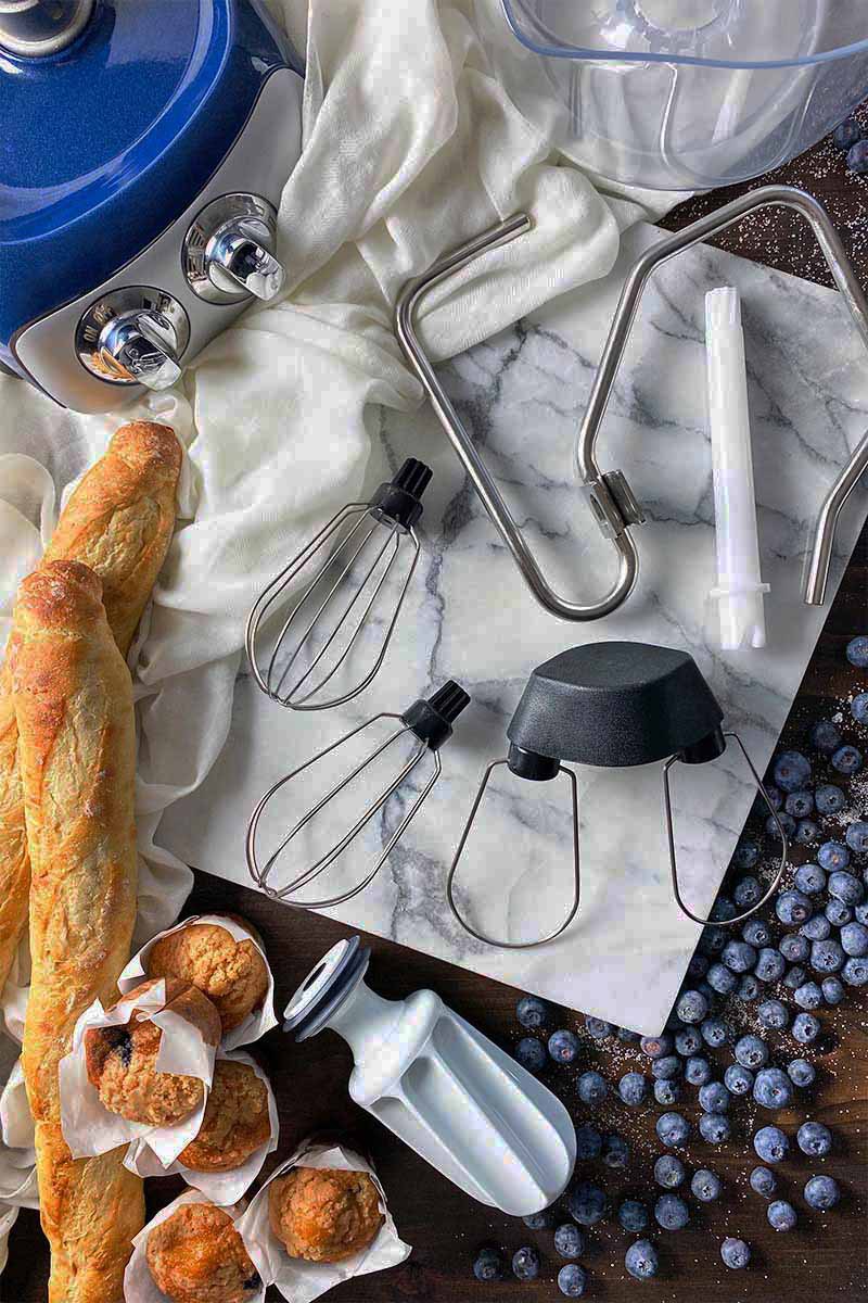Vertical top-down image of various tools and attachments on a marble board next to a mixer base, bread, muffins, and blueberries.