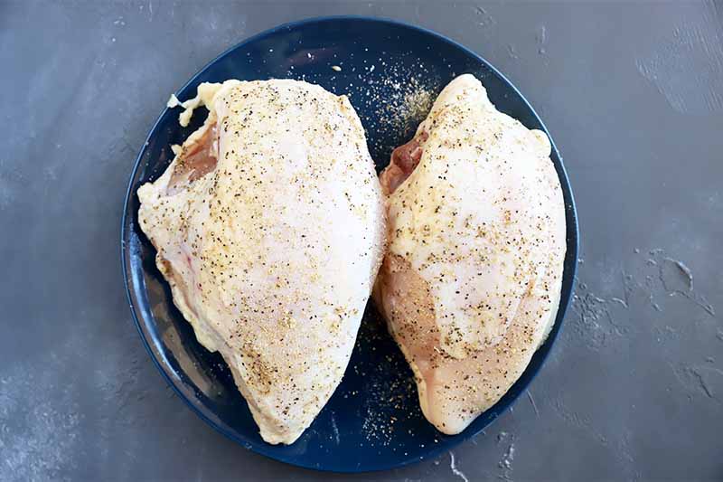 Horizontal image of two raw poultry pieces seasoned with salt, pepper, and spices on a plate.
