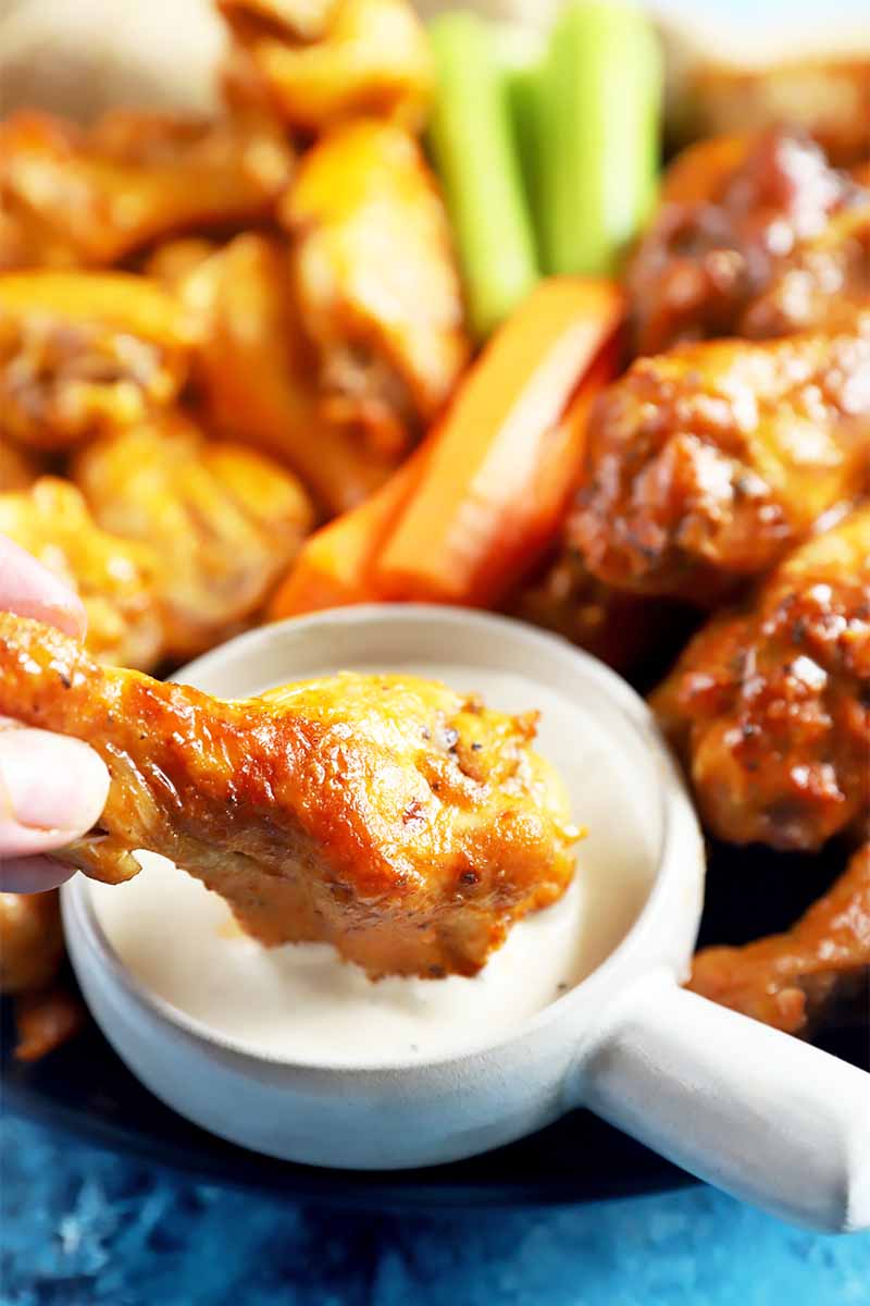 Vertical image of a hand dipping a drumstick in white dressing next to celery sticks and carrot sticks on a blue surface.