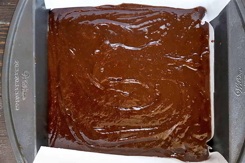 Horizontal image of raw thick cocoa batter in a square pan lined with parchment paper.