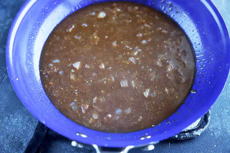 Horizontal image of a blue pan cooking a brown sauce with chopped onions.