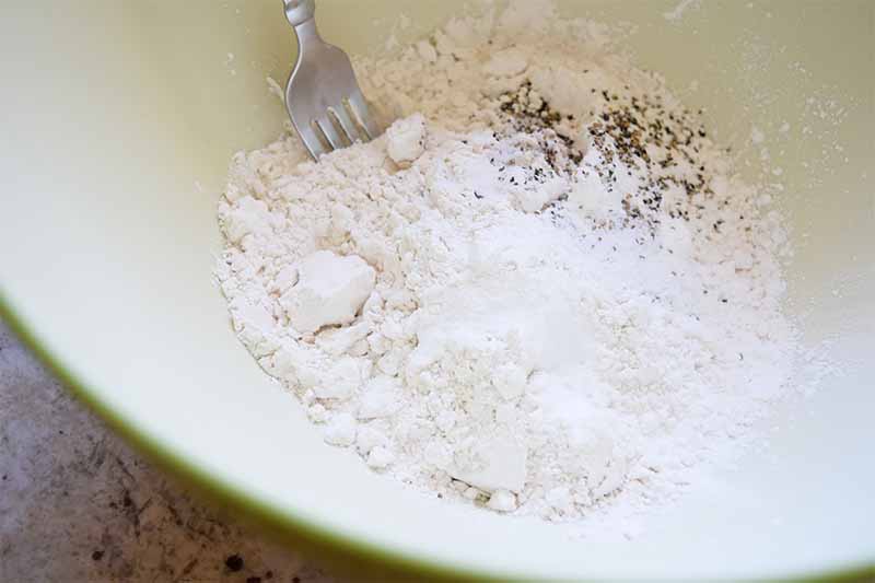 Horizontal image of dry ingredients in a bowl mixed by a metal fork.