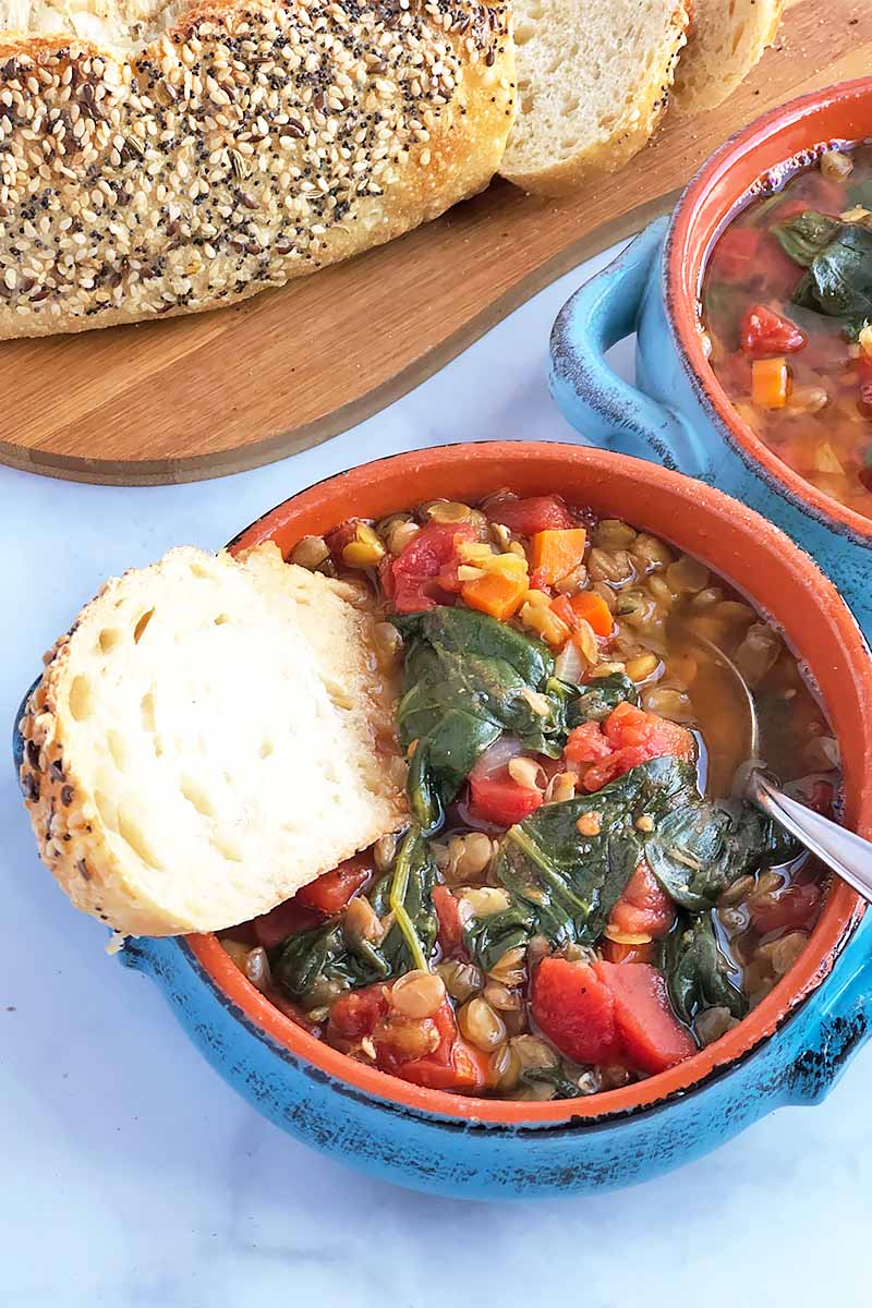 Vertical close-up image of a bowl filled with a vegetable stew with a metal spoon and a slice of bread, with more bread on a wooden cutting board in the background.