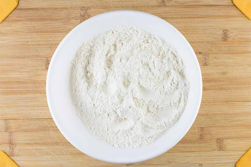 Horizontal image of a white bowl filled with sifted dry ingredients on a wooden cutting board.