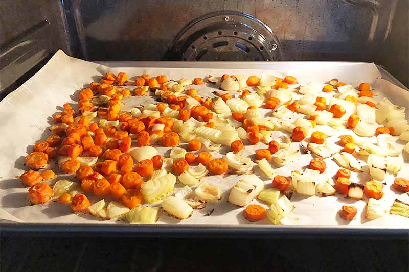 Horizontal image of roasting chopped carrots and onions on a lined baking sheet.