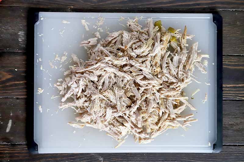 Horizontal image of shredded cooked meat on a white cutting board.