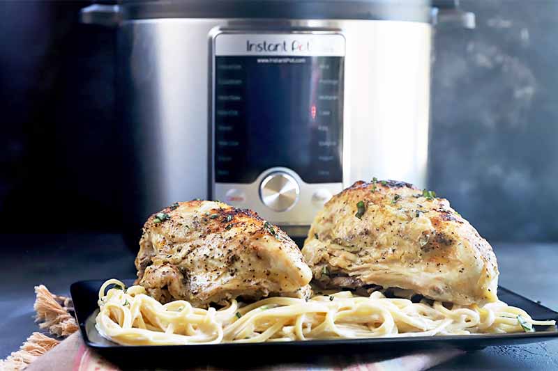 Horizontal image of two cooked piece of poultry with seasonings on top of spaghetti on a black plate in front of an appliance.