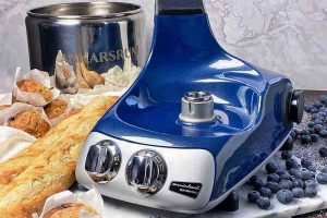 Horizontal image of the base of a blue mixer next to a metal bowl, bread, muffins, and blueberries.