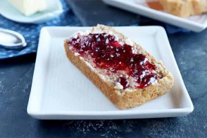 Horizontal image of a slice of bread covered in butter and jam on a white square plate.