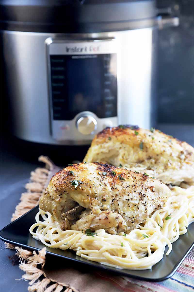 Vertical image of of pieces of cooked and seasoned poultry on a plate of spaghetti in front of a large kitchen appliance.