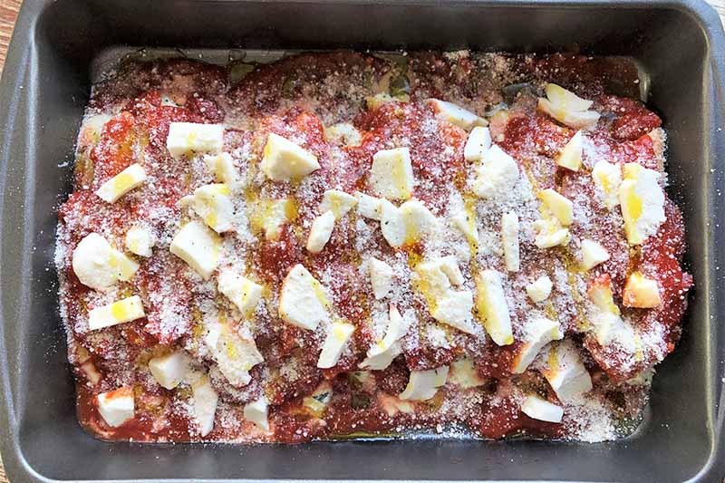 Horizontal image of an unbaked casserole covered with marinara and assorted cheeses in a pan.