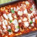 Horizontal image of a rectangular metal baking dish filled with a casserole completely covered in tomato sauce, herbs, and melted mozzarella.