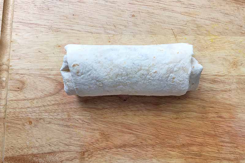 Horizontal image of a neatly rolled burrito on a wooden cutting board.