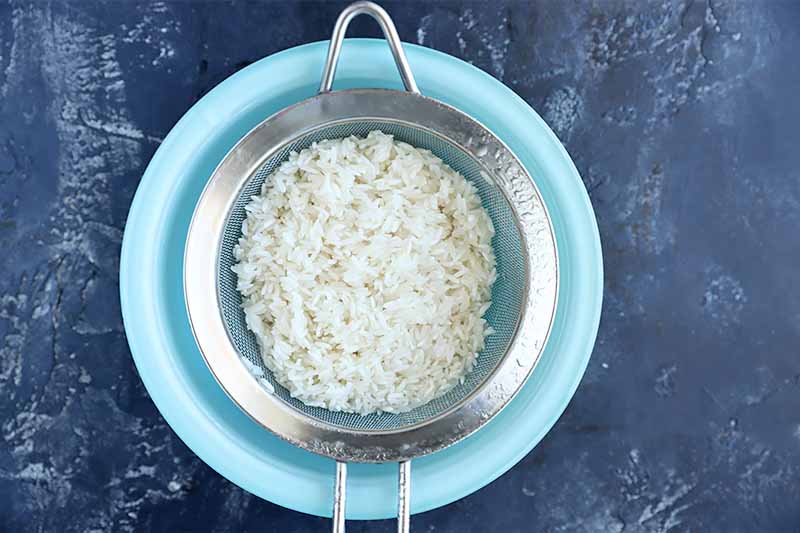 Horizontal image of rinsing rice in a strainer over a blue bowl.
