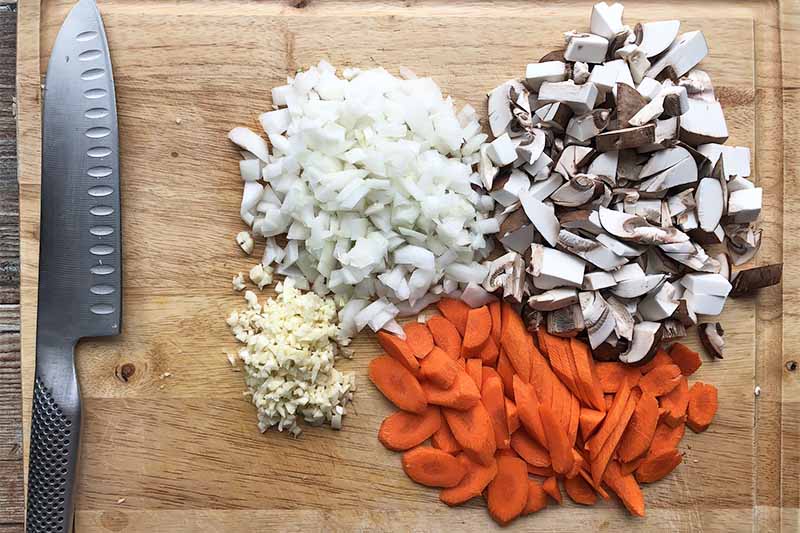 Horizontal image of chopped carrots, mushrooms, onions, and garlic on a wooden cutting board next to a knife.