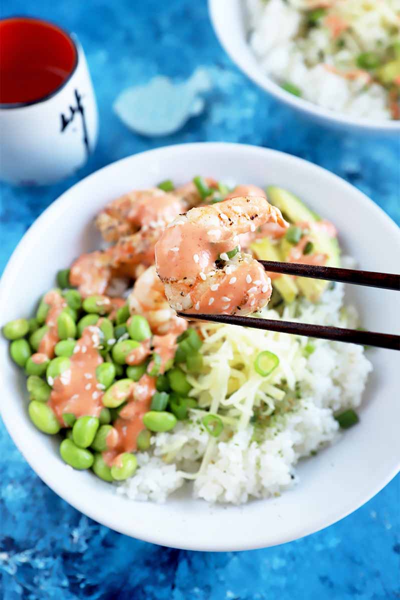 Vertical image of chopsticks holding a piece of shrimp over a white dish with rice and edamame on a blue surface.