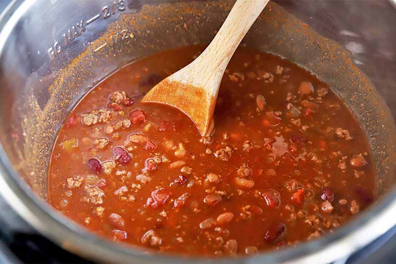 Horizontal image of a wooden spoon stirring a thick chili mixture in a pot.