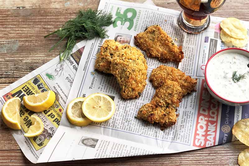 Horizontal image of three pieces of breaded and baked fillets on a newspaper next to lemons, herbs, and a bowl of a creamy white dressing.