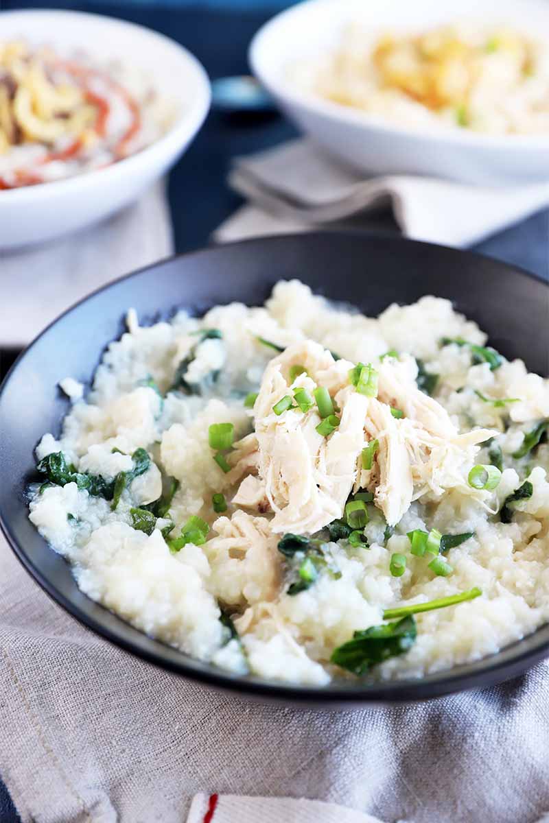 Vertical image of a black bowl filled with a thick porridge topped with greens and shredded chicken.