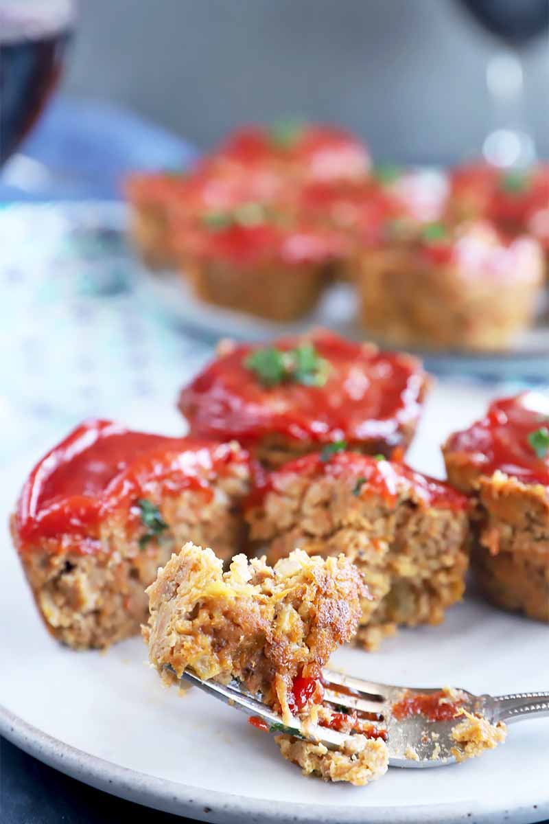 Vertical image of a fork taking pieces from small meatloaf muffins topped with ketchup.