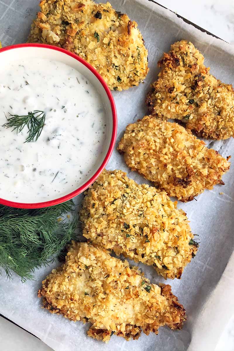 Vertical image of a baking sheet lined with parchment paper, with five pieces of breaded and baked meat on top next to a bowl full of a creamy white dressing and herb garnish.