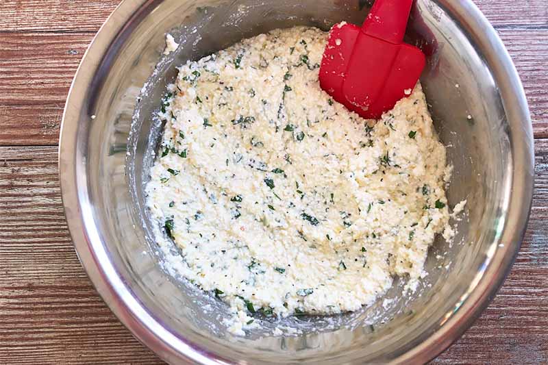 Horizontal image of a ricotta and herb mixture in a metal bowl being stirred by a spoon.