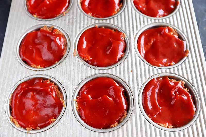 Horizontal image of a muffin pan filled with a mixture topped with a thick layer of ketchup.
