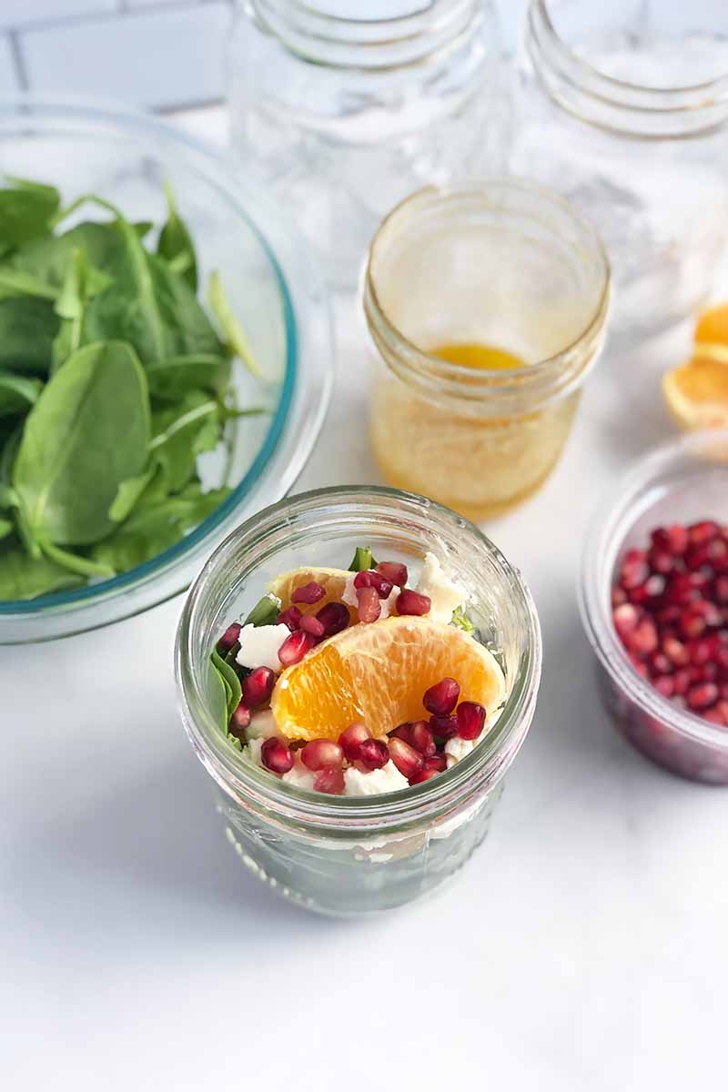 Vertical image of a portioned salad in a glass bowl next to a bowl of spinach, dressing, pomegranate seeds, and orange slices.