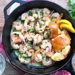Horizontal image of a cast iron skillet filled with cooked seafood in a citrus sauce next to chunks of baguette and lemon slices on a wooden table surrounded by the same ingredients.