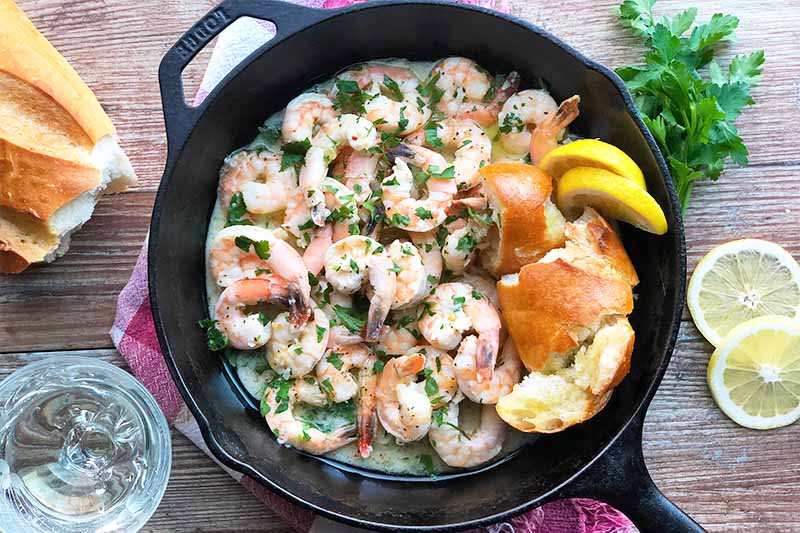 Horizontal image of a cast iron skillet filled with cooked seafood in a citrus sauce next to chunks of baguette and lemon slices on a wooden table surrounded by the same ingredients.