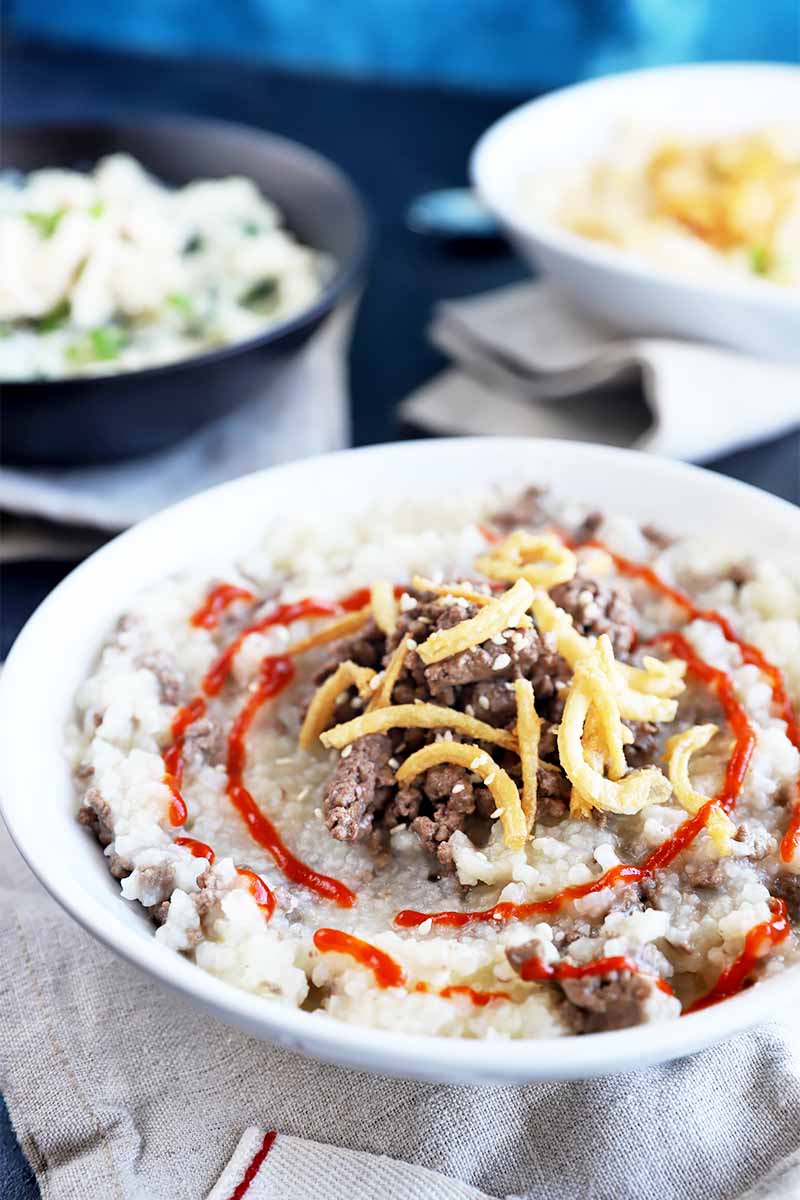 Vertical image of a white bowl filled with a thick porridge topped with meat, fried onions, and red sauce drizzle.
