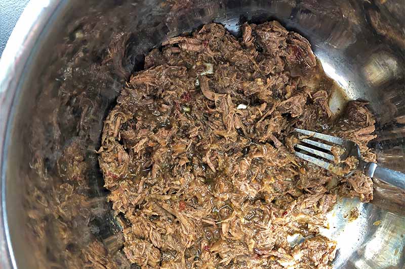 Horizontal image of shredded meat and a metal fork in a pan.