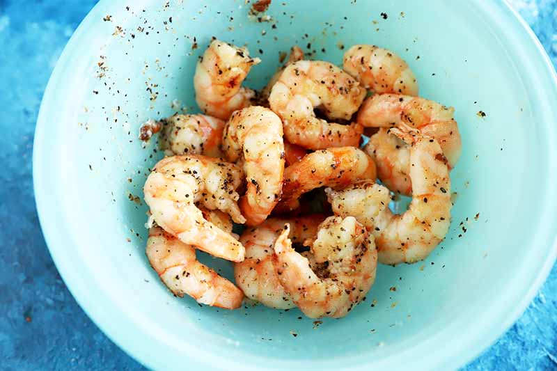 Horizontal image of a blue container with seasoned cooked shrimp tossed inside.