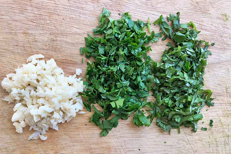 Horizontal image of minced garlic and herbs on a wooden cutting board.