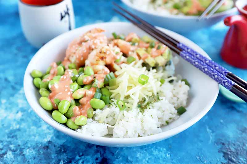 Horizontal image of a white dish filled with cooked grains, edamame, cooked seafood and a drizzle of a creamy red sauce next to chopsticks on a bright blue surface.