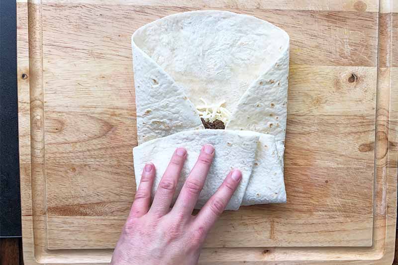 Horizontal image of a hand folding a large flour tortilla on a wooden cutting board.