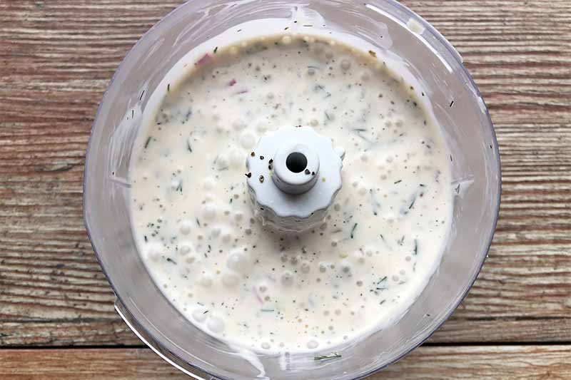 Horizontal image of a food processor filled with a thick white liquid seasoned with fresh herbs.