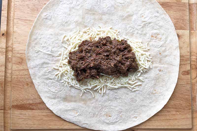 Horizontal image a pile of shredded cooked meat and shredded cheese in the center of a large flour tortilla.