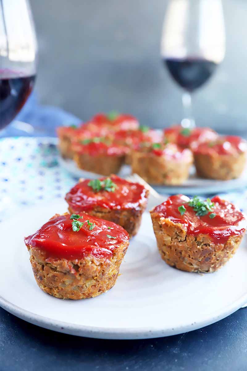 Vertical image of scattered mini beef muffins topped with ketchup and herbs on a plate in front of a blue napkin and glasses of red wine.