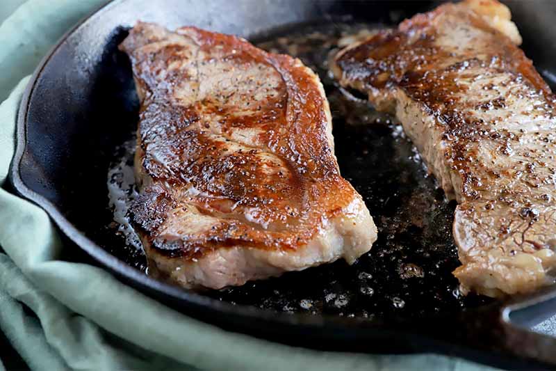 Horizontal close-up image of two thick pieces of meat browned and seasoned in an oiled cast iron.