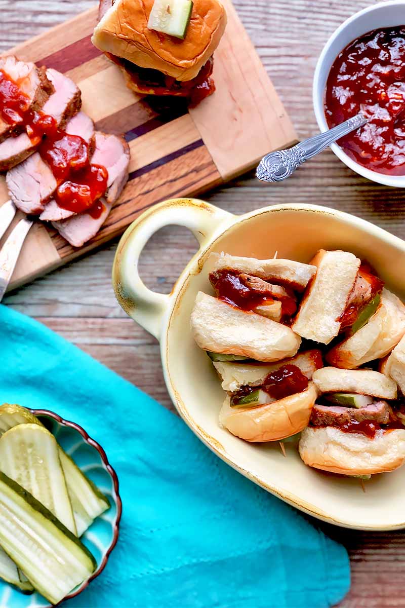 Vertical top-down image of sliced meat covered in sauce on a wooden platter next to mini sandwiches in a white tray next to a blue towel with a bowl of sliced pickles on top and a bowl of a red condiment with a spoon.